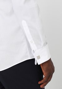 Hvit skjortekrage med jevn tekstur, med firkantede mansjettknapper og knappeåpning på mansjetten. Stoffet har en mild glans.