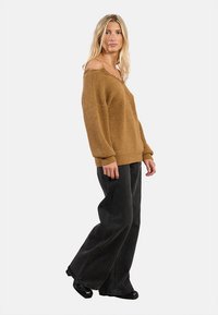 Maglione in maglia marrone oversize con trama testurizzata, abbinato a jeans neri a gamba larga; design rilassato e stile scivolato sulle spalle.