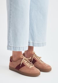 Piedi che indossano sneakers in suede beige e bordeaux con lacci beige, abbinati a jeans in denim a vita corta di colore blu chiaro, su uno sfondo bianco.