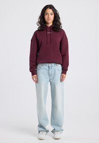 Woman standing facing forward wearing a maroon hoodie, light blue loose jeans, and white sneakers on a plain white background.