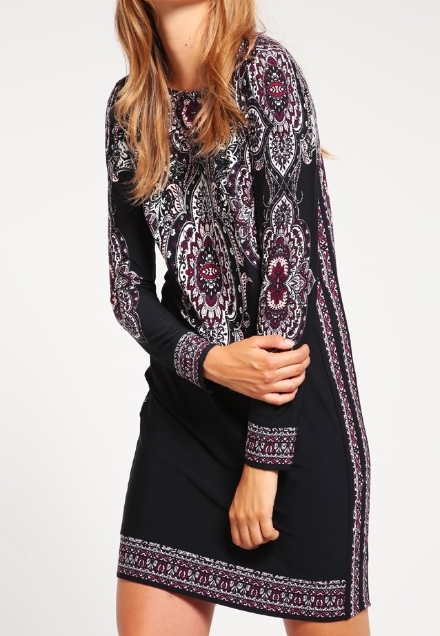 Femme portant une robe noire avec des motifs en paisley intriqués en violet et blanc sur les manches, le devant, les poignets et l'ourlet, se tenant les bras croisés.