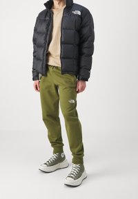 Person som bär svart pufferjacka, beige skjorta, olivgröna joggers och gröna högskor står mot en enfärgad vit bakgrund.