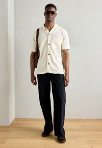 Cammicia a maniche corte in maglia crema con bottoni neri e tasca, abbinata a pantaloni a gamba larga blu scuro e mocassini marroni.