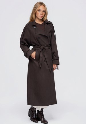 Un mannequin pose avec un trench croisé marron foncé ceinturé, porté sur un haut en maille, une jupe blanche et des bottines noires.