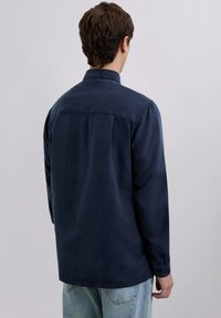 Camisa de manga larga azul marino de tela suave, con un cuello clásico y puños abotonados, vista desde la espalda contra un fondo claro.