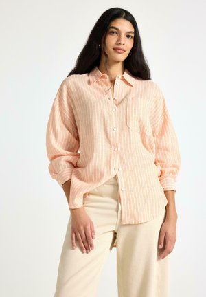 Donna in piedi che indossa una camicia a righe larghe pesca e bianca con bottoni, abbinata a pantaloni beige, su uno sfondo bianco semplice.