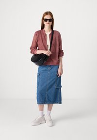 Růžová texturovaná bomber bunda, béžový top, modrá džínová midi sukně s kapsami a bílé tenisky se žlutými detaily. Černá taška a sluneční brýle.