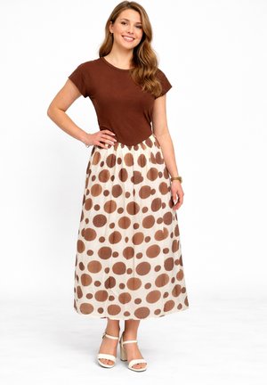 Lachende vrouw staat met hand op heup, draagt een bruin kort mouwtopje, witte rok met grote bruine stippen en witte sandalen met hak.