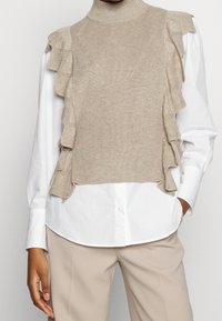 Beige neulottu liivimallinen neulepusero, jossa on korkea kaulus ja röyhelökoristeet hihojen ympärillä, pukeutuneena valkoisen napitettavan paidan päälle. Teksturoidulla pinnalla.