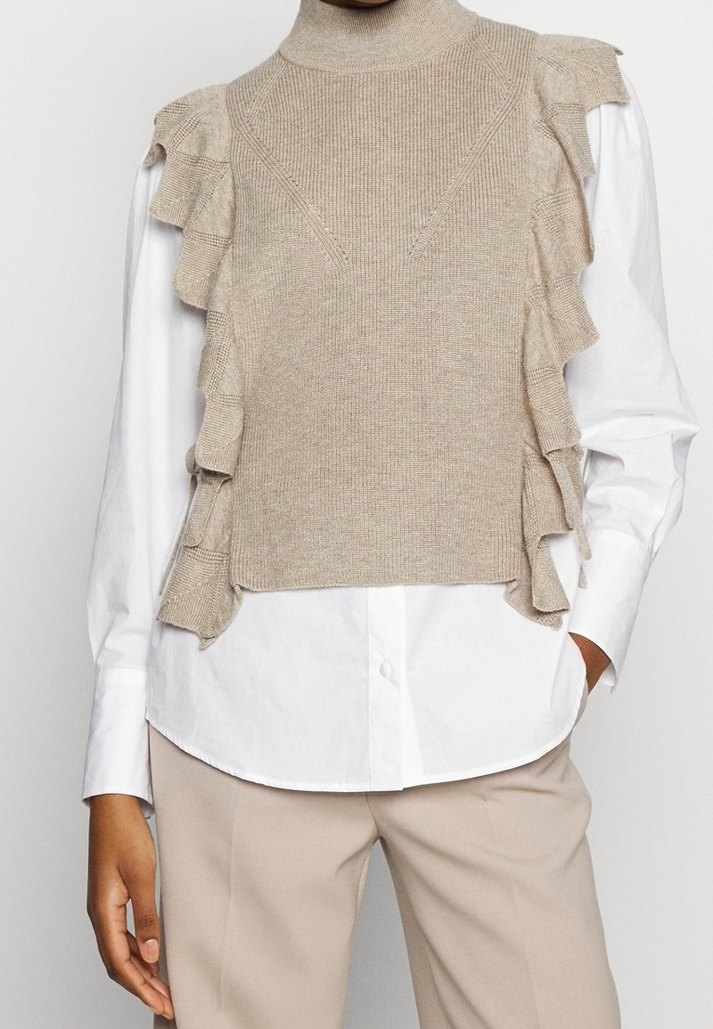 Beige neulottu liivimallinen neulepusero, jossa on korkea kaulus ja röyhelökoristeet hihojen ympärillä, pukeutuneena valkoisen napitettavan paidan päälle. Teksturoidulla pinnalla.