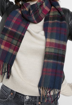 Personne portant une écharpe à carreaux multicolores sur un pull crème et une veste en cuir noire, visible des épaules aux hanches.