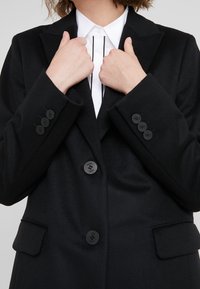 Person som justerar slagena på en svart knapplös blazer över en vit skjorta med svarta vertikala ränder.