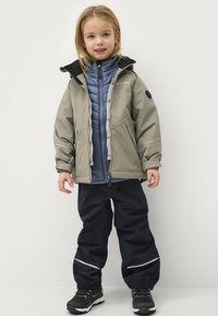 Kind in een grijze waterdichte jas, blauwe geïsoleerde binnenjas, zwarte broek en zwarte schoenen met reflecterende accenten.