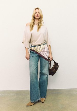 Kvinde iført off-shoulder beige top, vidde ben blå jeans, spidse brune sko, holder en brun taske, med et stribet tørklæde bundet om taljen.