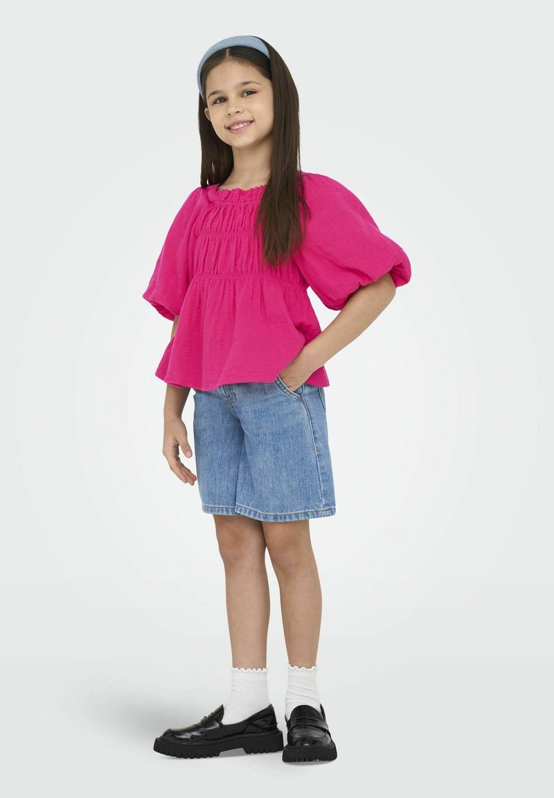 Lys pink kortærmet bluse med samlede detaljer, sat sammen med blå denim shorts og sorte loafers. Hvide bølgede sokker fuldender outfittet.