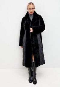 Zwarte nepbont en gewatteerde stof jas, half lang, met grote kraag, knopen aan de voorkant en zijsplitten. Draagt met zwarte laarzen tot de knie.