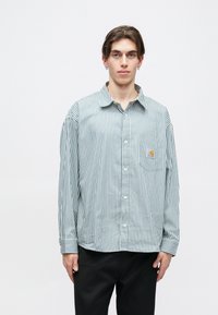 Camisa de botón a rayas en verde y blanco, con bolsillo en el pecho y etiqueta de marca, confeccionada con tela ligera y corte relajado.