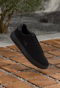 Zwarte sneaker met een gladde textuur, heeft tonal branding, platte veters en een dikke rubberen zool, geplaatst op een gestructureerde stenen ondergrond.
