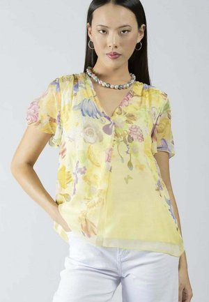 Donna che indossa una blusa a maniche corte gialla a fiori, pantaloni bianchi, orecchini a cerchio e una collana di perline, in posa con una mano in tasca.