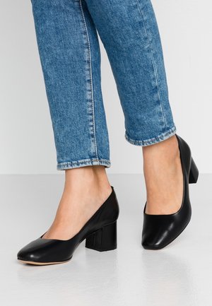 Pieds portant des chaussures noires à talons blocs associées à un jean denim bleu raccourci au-dessus des chevilles sur un fond uni.