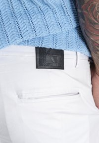 Blauer Strickpullover über weißen Hosen, mit einem schwarzen Lederdetail auf dem Bund, das die Initialen "KG" trägt, und einer Rückentasche.