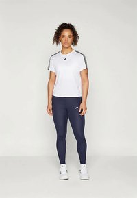 Vit idrottströja med svarta ränder på axlarna, tillsammans med marinblå leggings, som båda har Adidas-logotypen.