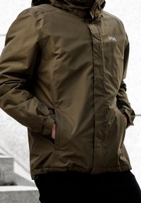 Personne portant une veste imperméable Helly Hansen olive avec les mains dans les poches, sur un fond extérieur flou.