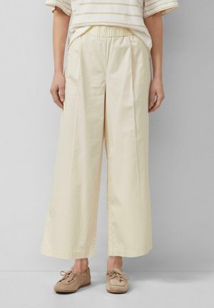 Frau trägt cremefarbene, weit geschnittene Hose mit elastischem Bund und beige Mokassin-Schuhe, steht vor einem schlichten grauen Hintergrund.