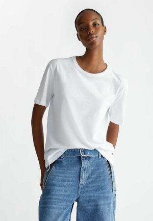 T-shirt bianco in cotone con scollo rotondo e maniche corte, abbinato a jeans in denim blu con dettagli a zip sui lati.