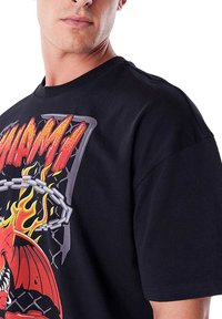 T-shirt nera in cotone con una grafica colorata che presenta fiamme, una catena e la parola "MIAMI" in lettere rosse in grassetto. Maniche corte, vestibilità rilassata.