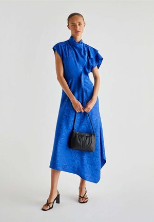 Asymmetrische blauwe jurk van glanzend materiaal met korte mouwen en textuurpatronen. Gecombineerd met een zwarte handtas en sandalen met blokhakken.