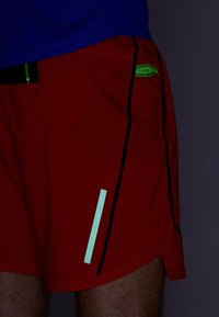 Shorts de sport rouges avec des coutures latérales noires, une bande verticale vert menthe et un accent de poche vert contrastant. Tissu léger.