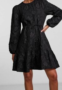 Svart texturerad blommig klänning med långa puffärmar, knytning i midjan och volangkant, bärs av en stående person.