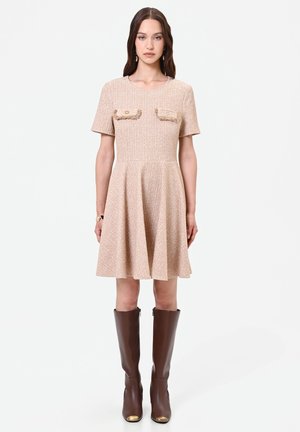 Donna che indossa un vestito beige a maniche corte con due tasche sul petto, abbinato a stivali di pelle marroni al ginocchio.