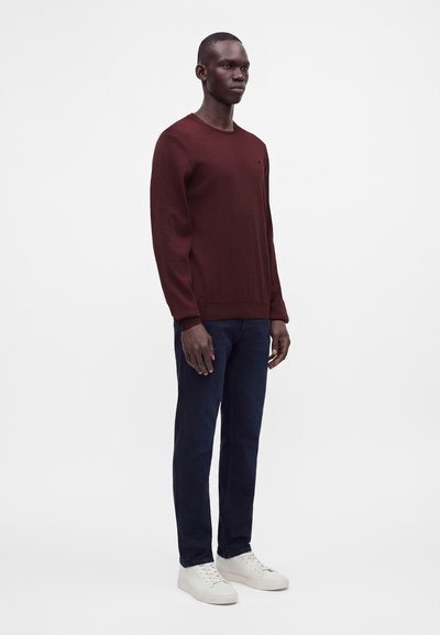 Homme debout portant un pull rouge foncé, un pantalon bleu marine et des baskets blanches sur un fond blanc uni.
