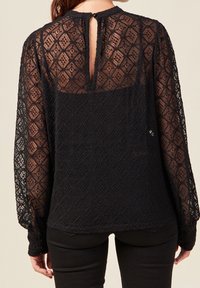 Blusa nera in pizzo con maniche lunghe, motivo a diamante, chiusura a chiave sul retro e polsini elasticizzati. Il tessuto trasparente e testurizzato aggiunge profondità.