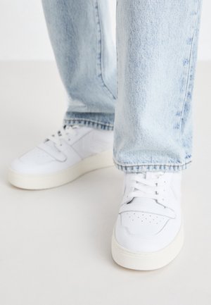 Persona con jeans azul claro y zapatillas blancas de pie sobre una superficie lisa y clara, con vistas frontales y laterales de los zapatos visibles.