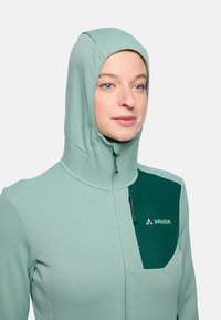 Felpa zip-up verde chiaro realizzata in materiale morbido, con cappuccio aderente, tasca sul petto verde scuro e chiusura a zip.