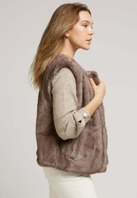 Femme aux longs cheveux blonds portant un gilet en fausse fourrure taupe sur un pull beige et un pantalon blanc, posée de profil sur fond uni.