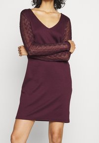 Šaty s výstřihem do V v barvě burgundy s dlouhými krajkovými rukávy, přiléhavý střih a jemná textilie; délka do poloviny stehna s hladkou sukní.