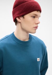 Felpa color teal con collo a costine e patch con logo ricamato. Il modello indossa un berretto rosso a costine. Texture morbida, design minimalista.