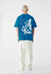 Blå t-shirt med korte ærmer, der har et hvidt abstrakt design på ryggen, parret med strukturerede hvide bukser og hvide sneakers.