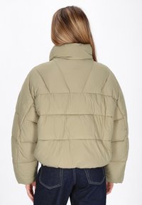Donna con capelli lunghi che indossa una giacca imbottita verde oliva chiaro e jeans blu scuro, vista di spalle su uno sfondo bianco.