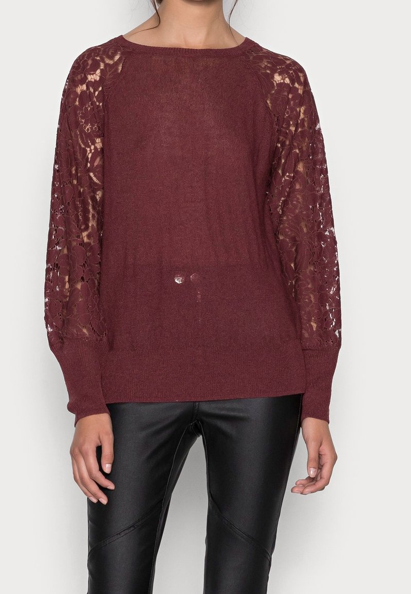 Femme portant un pull en maille bordeaux avec des manches longues en dentelle à motif floral et un pantalon en cuir noir, debout devant un fond blanc.