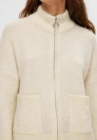 Crème-kleurige cardigan van zacht gebreid materiaal. Heeft een hoge kraag, een rits aan de voorkant en twee voorzakken met ribbelranden.