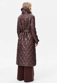 Brązowy pikowany długi płaszcz z wysokim kołnierzem, wiązanym w talii i detalami z logo. Posiada błyszczące wykończenie i skośny wzór przeszyć.