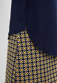 Tmavě modrý top přeskládávající se s černou, bílou a žlutou vzorovanou sukní s geometrickými diamantoidními a čtvercovými tvary.