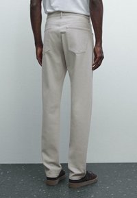 Pantaloni in cotone beige con un design a gamba dritta, dotati di due tasche posteriori e vita a media altezza. Indossati con scarpe scure.