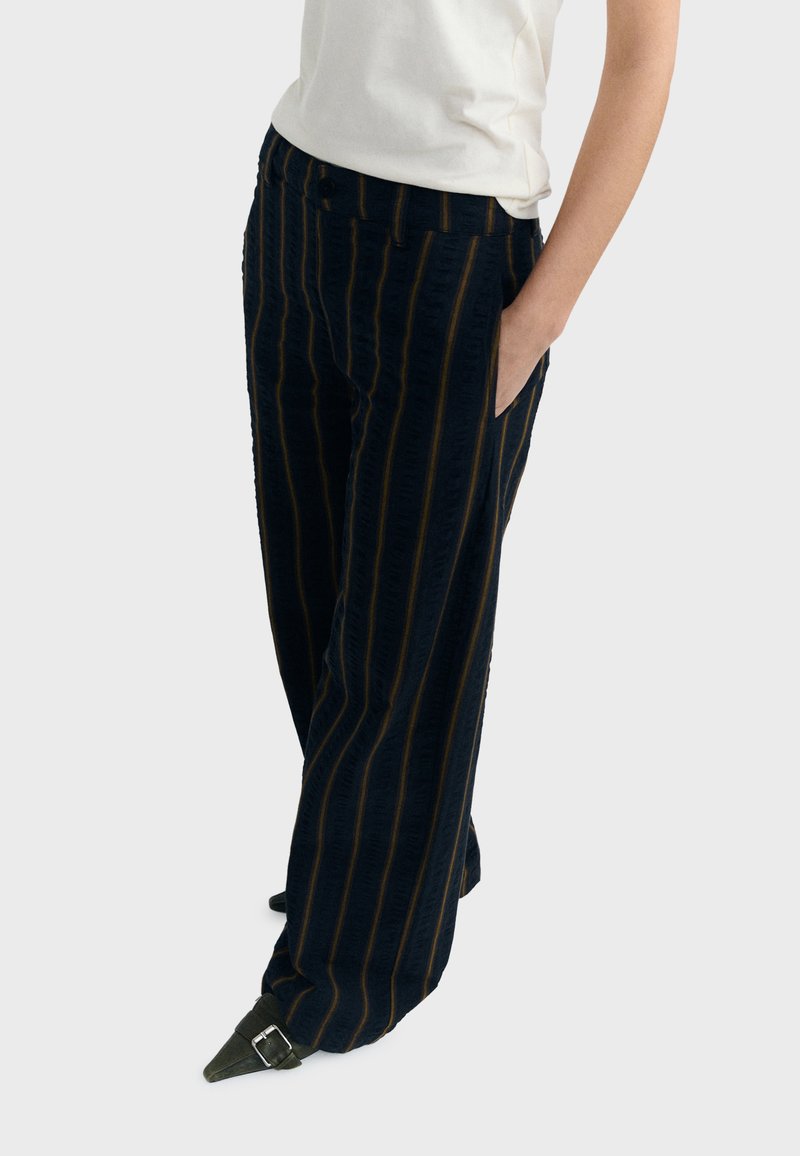 Personne portant un pantalon large noir à rayures verticales marron, un t-shirt blanc et des chaussures pointues foncées avec une boucle, main dans la poche.