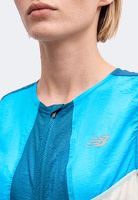 Близък план на човек, носещ син спортен суичър с цип и лого на New Balance на гърдите.
